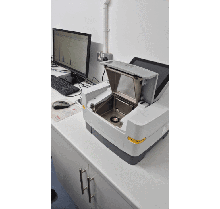 A laboratory bench with a desktop computer and an open analytical instrument, likely a spectrometer or X-ray machine, revealing its sample compartment. The setting is clean and organised.