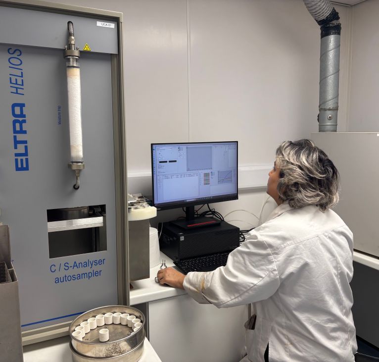 Technician in a lab coat using an ELTRA HELIOS C/S elemental analyzer autosampler while reviewing carbon and sulfur analysis data on a computer monitor.