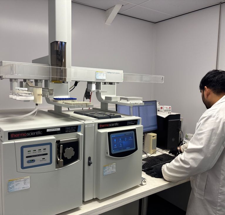Lab technician in a white coat using a Thermo Scientific gas chromatography tandem mass spectrometry (GC–MS/MS) instrument with an automated autosampler and computer.
